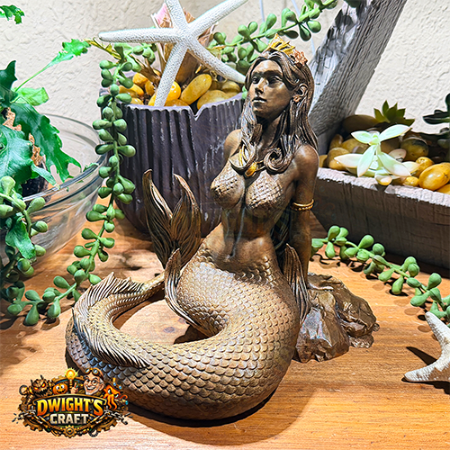 A bronze-finished mermaid figurine with a crown and gold jewelry, styled on a wooden surface with ocean-themed decor including a starfish, green trailing plants.