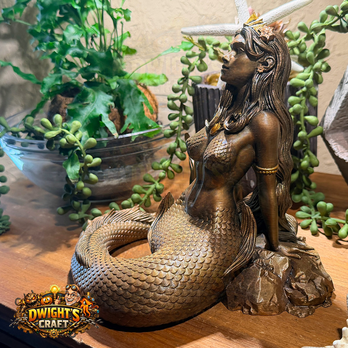 A bronze-finished mermaid statue with a detailed scale tail, seated on a rock base with a starfish fin, displayed on a wooden surface with green plants in the background.