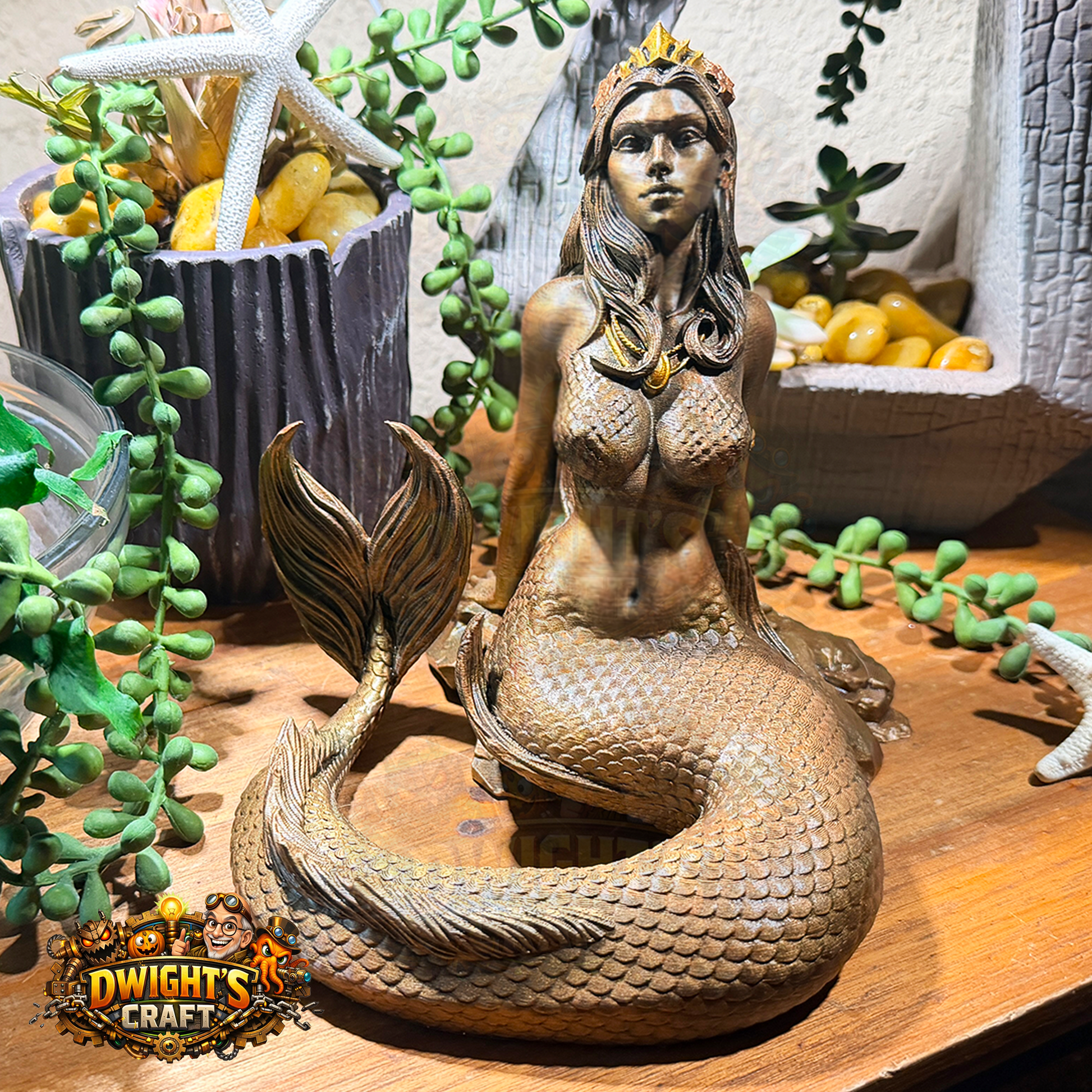 A bronze-finished mermaid figurine with a crown and pearl necklace, displayed on a wooden surface with decorative plants and a pot in the background.