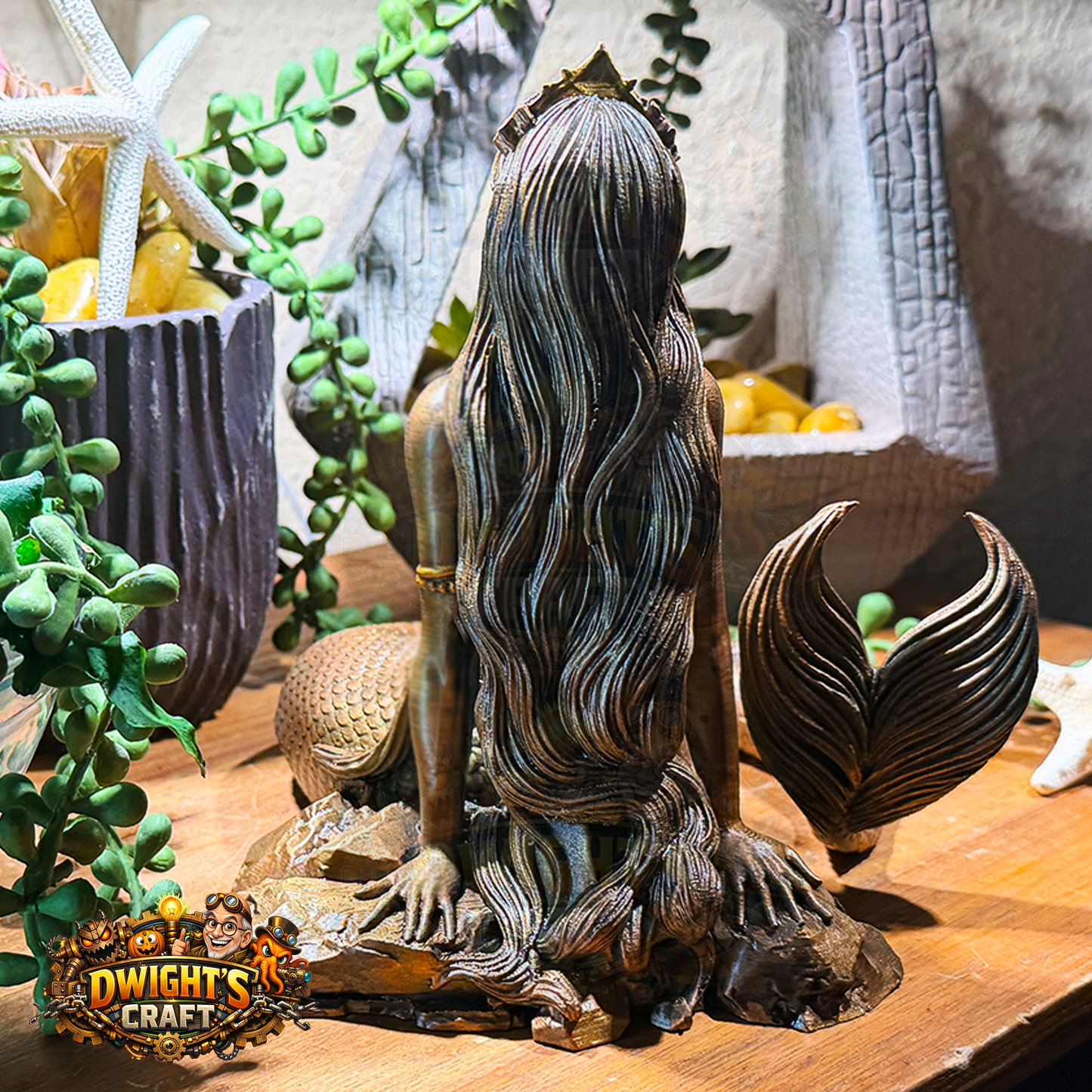 A bronze-finished mermaid statue shown from the back, seated on a rocky base with long flowing hair and a detailed tail, displayed on a wooden surface.