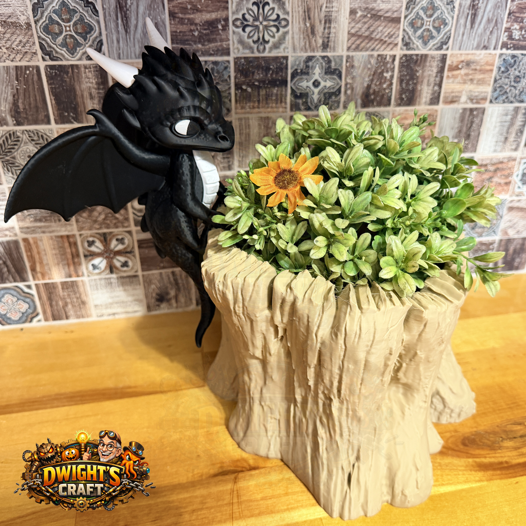A black dragon figurine with white horns and wings stands beside a faux green bush in a tree-stump planter on a wooden surface, with a tiled wall behind it. A 'Dwight's Craft' logo is visible in the lower-left corner.