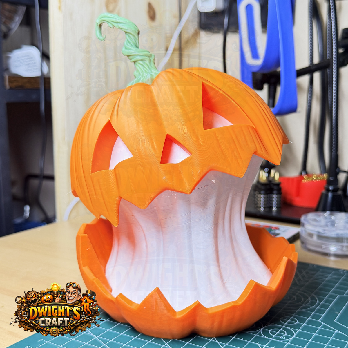 A 3D-printed jack-o'-lantern pumpkin with a carved face and open mouth, sitting on a green cutting mat in a workshop setting. A 'Dwight's Craft' logo is visible in the bottom-left corner.