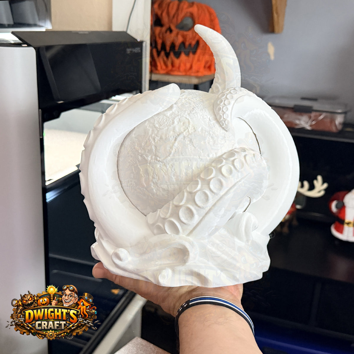 A hand holds a white, unpainted 3D-printed pumpkin-shaped ornament with tentacles wrapping around it, set against a cluttered indoor background with a 3D printer and Halloween decorations.