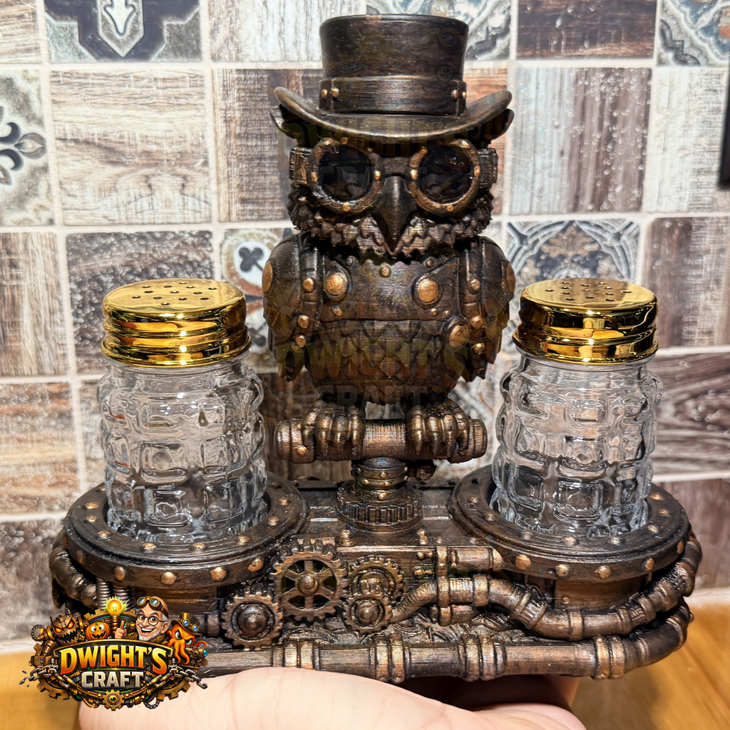 Steampunk owl salt and pepper shaker set with a bronze-finished owl on a gear-tracked base, flanked by two clear glass mason-jar shakers with gold lids, held in a hand against a tiled wall.