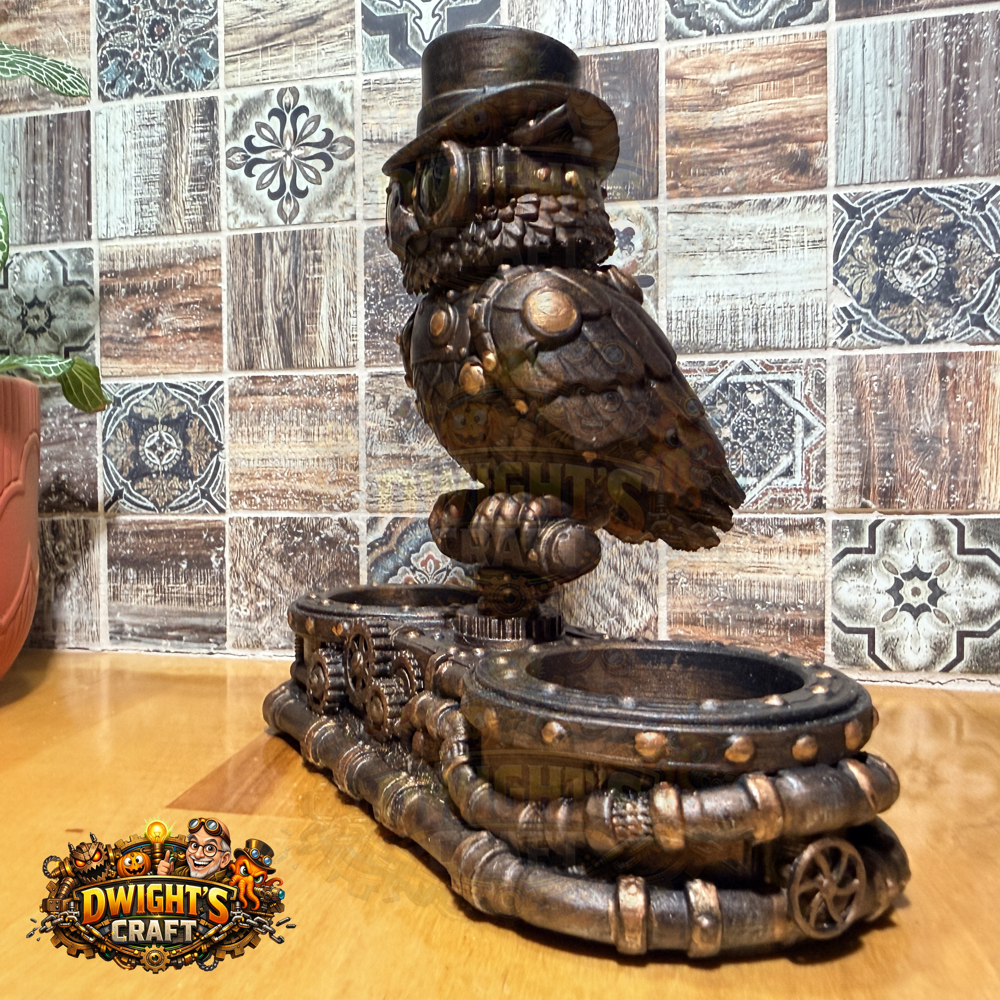 Steampunk owl candle holder and drink set on a wooden tabletop in front of a tiled backsplash. The owl figurine wears a small top hat and metallic armor details, perched on a pipe-like base with two cup holders.